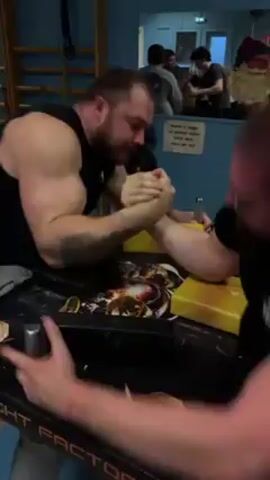 Two men are arm wrestling at a table. The man on the left is muscular and has a beard and tattoos on his left arm. The man on the right wears glasses.  There is a yellow block and other objects on the table. Other people are in the background.