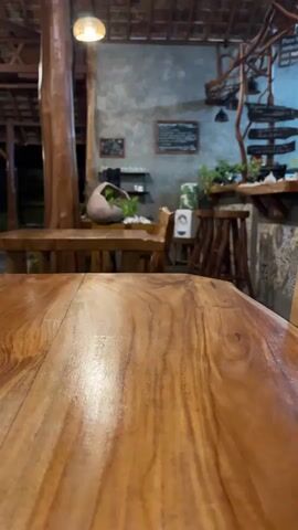 This is a medium shot of a wooden table in the foreground, with a blurred background of a rustic-style cafe or restaurant. The cafe has a thatched roof, wooden pillars and furniture, and concrete walls. There are plants on shelves and around the seating area, giving it a natural, earthy feel. A light fixture hangs from the ceiling, and there are signs on the wall. The image appears to be taken at dusk or in the evening, as the lighting is dim and artificial. The overall mood is relaxed and inviting. There are no people visible in the frame. The focus is on the textures and warm tones of the wood, creating a cozy and natural ambiance.
