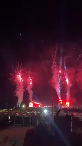 The video shows a dark nighttime scene with bright red and blue pyrotechnics firing into the sky. In the foreground, there are tents and what appears to be a parking area with a white van. In the background, there is a large stage with multiple pillars of light and smoke rising from it. The pyrotechnics create bursts of light and smoke that illuminate the night sky. The overall atmosphere is lively and festive, suggesting a concert or special event.