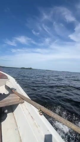 The video shows a view from a white boat on a body of water under a partly cloudy blue sky. A wooden oar is visible in the foreground, extending from the right side of the boat and angled towards the left. Water splashes behind the oar, indicating the boat is moving. The horizon shows a distant shoreline with some greenery and a few buildings. The sky has wispy white clouds. The video appears to be a first-person perspective from someone in the boat.