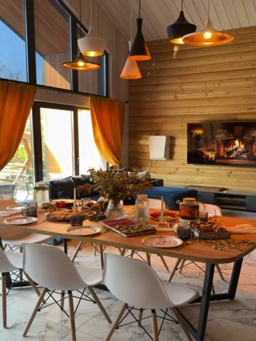 The video shows a dining room in a house with wooden walls. The room features a long wooden table set for a meal, surrounded by white chairs. The table is decorated with food, drinks, cutlery, plates, candles, and a floral arrangement in a vase. A blue couch is situated near a large window covered with yellow curtains. Pendant lights hang above the table. A television mounted on the wooden wall displays a fireplace scene. The floor is covered with marble-patterned tiles. The video focuses on the details of the dining arrangement.