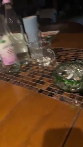 The video shows a close-up of a tiled surface with a green object on the left side of the frame. The tiling is made of small, square mosaic tiles in various shades of brown and black, with a band of lighter cream and beige tiles in the middle. The green object appears to be a glass, possibly a wine glass or a vase, and it is out of focus. The background is dark and blurry, suggesting it might be nighttime or an indoor setting with low lighting. There is a brief audio clip at the beginning, which appears to be a voice asking a question. The video pans slightly to the right, showing more of the tiled surface and the blurry background.