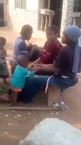 The video shows four people outdoors in front of a patterned building. Three women are seated on a concrete block. The woman to the right has a blue and grey striped head covering and wears a black top and jeans. The woman in the middle has a maroon shirt and jeans and is talking. The woman to the left has a striped shirt. A young child stands between the women. Another child is partially visible to the left. The ground is dirt with scattered debris. A white object is in the foreground. The woman in the middle gestures and drinks from a small bottle. The scene appears informal and domestic.