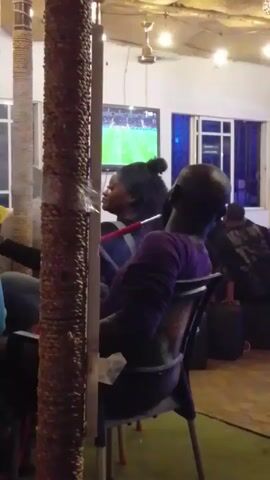 The video shows people sitting in a casual indoor setting, possibly a lounge or bar, with a TV playing what appears to be a soccer game in the background. Several people are seated, including one individual in a purple shirt who is turned away from the camera, and another woman with her hair in a bun. A hookah with a red hose is visible between them. The room is decorated with textured pillars, and has a ceiling with a thatched or woven design. There are windows letting in natural light and some pictures on the wall. The overall atmosphere seems relaxed and informal.