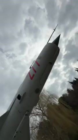 The video shows a static display of a gray MiG-21 fighter jet. The jet is mounted on a concrete pedestal in what appears to be a park or open area. The underside of the jet, including the wings and mounted missiles, is visible. The number "211" is painted in red on the fuselage. The sky is overcast with gray clouds. There are trees in the background and a person walking in the distance.