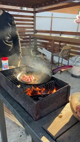 Here's a detailed description of the video:

The video shows a person cooking meat patties in a frying pan over an open fire on an outdoor grill. The grill is made of dark grey stone and sits on a wooden table. Inside, the fire is burning with orange and red flames. The frying pan, with a silver cooking surface and a red handle, rests on the grill grates.

A person, wearing a light green fleece jacket, is placing a raw meat patty into the frying pan. They're wearing a clear plastic glove on their right hand. Steam rises from the pan as the meat cooks. The meat patties are irregularly shaped and have a reddish color.

In the background, there is a wooden fence, some stacked white PVC pipes, and a wooden chair. Sunlight is filtering through, creating a bright, slightly hazy atmosphere.