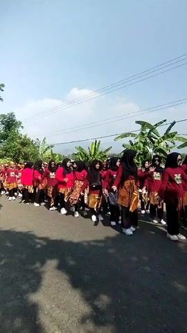 A group of women in matching red shirts and patterned skirts walk in a line outdoors on a sunny day. They are wearing headscarves and white shoes. Banana trees and foliage are visible in the background. The women appear to be participating in a parade or procession.