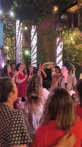 A group of women are dancing in a dimly lit outdoor venue. The lighting is dynamic, shifting between green, white, and purple hues. Large, vertical LED screens display abstract patterns of circles and lines. Green foliage hangs from above, and string lights are visible in the background. The women are dressed in various casual and semi-formal attire, including dresses and tops. They appear to be engaged in a celebratory event, possibly a wedding or a party, with some individuals holding hands and dancing in a circle. The overall atmosphere is lively and festive.