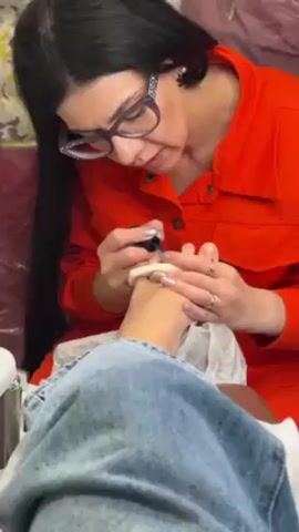 Here's a description of the video:

A woman in an orange jumpsuit and glasses is giving a pedicure to a person sitting in front of her. The person receiving the pedicure is wearing blue jeans and has their foot resting on a white, cushioned footrest. The woman is carefully applying nail polish to the toenails. The environment appears to be a nail salon or a similar beauty service setting.