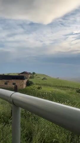 A wide shot captures a rural landscape under a cloudy sky. In the foreground, a silver pipe runs horizontally across the bottom of the frame, partially obscuring a chain-link fence. Beyond the fence, a lush green hillside is dotted with scattered trees and shrubs. Two small, weathered stone buildings with dark roofs are visible on the left side of the hill. The hillside slopes down towards a distant valley, where a town or city is faintly visible. The sky is a mix of blue and white clouds, with patches of sunlight breaking through, casting a warm glow on the horizon. The overall impression is one of a peaceful, natural setting with signs of human habitation.