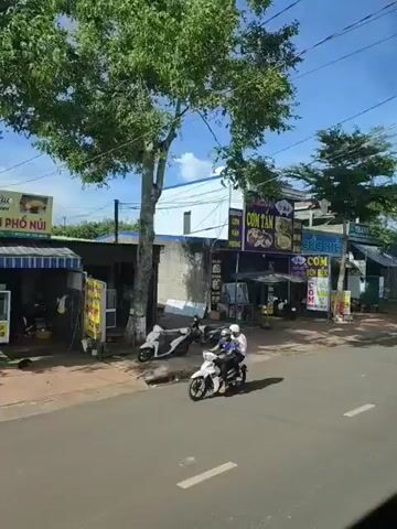 The video pans across a street scene with several businesses. The buildings are low-rise, likely one or two stories, with corrugated metal roofs in some areas. There are multiple signs in Vietnamese advertising food and services, including "CƠM TÂM," which translates to broken rice, a popular dish. Several motorbikes are parked in front of the establishments, and some people can be seen, though their actions are not clearly discernible due to the distance and camera movement. Trees line the street, and the sky is blue with some scattered clouds, indicating daytime. The overall impression is of a local commercial area in a Vietnamese town or city.