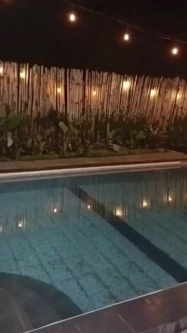 The video shows a swimming pool at night. The pool is rectangular and tiled in a blue and white pattern. String lights hang above the pool, and their reflections can be seen in the water. Behind the pool is a fence made of bamboo sticks. There are some plants growing along the base of the fence. The pool has a metal ladder leading into the water. The area around the pool is paved.