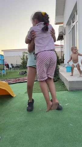 Two young girls are playing in a backyard. The girl in the foreground is wearing pink pajamas with a cloud pattern and checkered pink shorts. She is being held by another girl who is wearing a light blue t-shirt and shorts. The older girl has her arms around the younger girl. The younger girl is barefoot, and the older girl is wearing black Crocs. In the background, a toddler wearing a diaper is holding a blue bucket. There is a yellow slide to the left of the frame, a white house with an air conditioning unit, and a fence in the distance. The ground is covered with green turf. The sky is a pale orange, suggesting it is either sunrise or sunset.