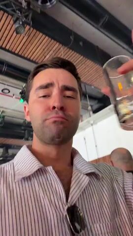 A man holds a drink in his right hand and films himself in a selfie video. He is in a dimly lit environment with green lighting. He is wearing a striped collared shirt. The camera movement is unsteady. The man smiles and makes a face at the camera. There are other people in the background, but they are blurred.