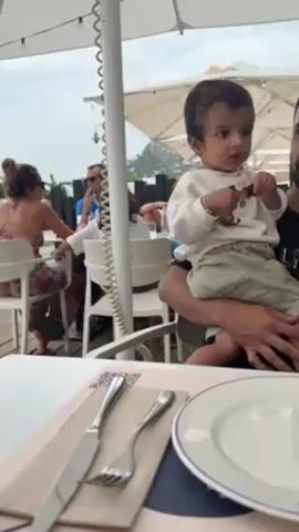 The video shows a restaurant scene. A baby sits on a man's lap at a table set with cutlery, a plate, and a menu. Another table is nearby with two women and a man sitting at it. One of the women is holding a phone. A cocktail is visible on the first table. The scene appears to be outdoors under umbrellas, with views of a road, trees, and a distant mountain.