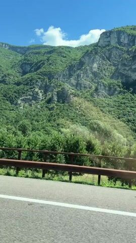 A car drives along a road with a metal guardrail on the right. Beyond the guardrail, there is a steep, green mountain covered in trees. The sky is blue with a few white clouds.