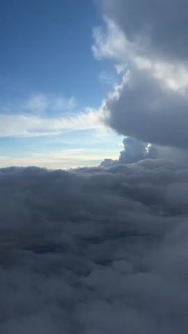 The video shows a view from above the clouds. The bottom of the frame is filled with a dense layer of dark gray clouds, resembling a thick blanket. Above this, there is a small section of a light blue sky with wispy white clouds. In the upper right corner, there is a large, puffy, gray cloud with some areas highlighted by sunlight.