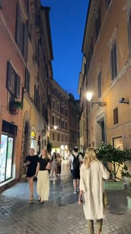 The video captures a lively evening scene on a narrow, cobbled street in Rome, Italy. Tall, multi-story buildings with warm-toned facades (shades of orange, yellow, and terracotta) line both sides of the street, their windows often adorned with dark, closed shutters and some featuring small balconies with plants. The sky above is a deep, clear blue of twilight, contrasting with the warm, golden glow emitted by ornate, wall-mounted streetlights that illuminate the bustling thoroughfare.

Numerous pedestrians, primarily young adults, are seen strolling along the cobblestone pavement. They are dressed in casual evening wear, walking in both directions, often in pairs or small groups. In the foreground on the left, two young women are smiling and conversing, one wearing a black top and a flowing white skirt, the other in a black top and shorts. A third woman in a light beige tunic and carrying a woven bag is visible from behind on the right, walking away from the camera. Other individuals can be seen throughout the mid-ground, some holding beverages or small items, contributing to the lively atmosphere.

The street-level areas feature illuminated shop windows, suggesting commercial activity. On the left, a brightly lit shop display is visible, while on the right, potted greenery hints at an establishment entrance. The overall mood is relaxed and pleasant, capturing the essence of an enjoyable evening walk through a historic European city. No discernible text is visible.