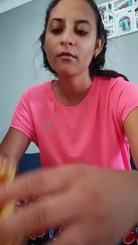A woman with dark hair pulled back wears a bright pink t-shirt and is seated at a table. Her hands are in the foreground, appearing to hold a yellow object. The background is out of focus, but hints at a room with a patterned surface on the table and what looks like a shelf with items on the wall.