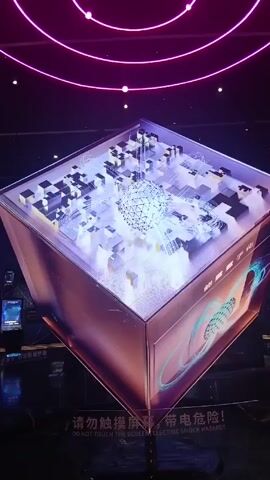 The video shows a lit-up, futuristic-looking cube exhibit. The cube is elevated, and above it are lit-up purple rings. The top of the cube appears to have a glass surface, under which is a white and black grid-like structure with a spherical object in the center. Light emanates from the sphere and flows through the grid. The side of the cube features text in Chinese and English, warning viewers not to touch the screen due to electric shock hazards. In the background, there are other screens and displays. The scene is darkly lit, with the main illumination coming from the cube itself and the rings above.