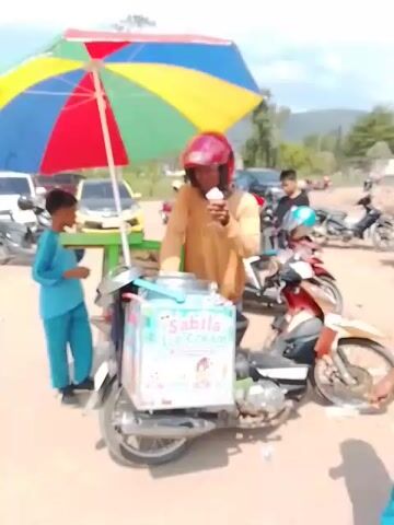 A man wearing a red helmet and an orange long-sleeved shirt is on a motorcycle that is outfitted with an ice cream cart. The ice cream cart is a light blue box with colorful graphics and the words "SABILA ICE CREAM" printed on it. The man is holding a small white cup with an ice cream cone in it, which he appears to be eating. A young boy in a blue tracksuit is standing to the left of the motorcycle, looking at the man. A large, colorful umbrella is propped up over the motorcycle and cart. The background shows a dirt road, parked cars, and motorcycles, and hills in the distance under a partly cloudy sky. The overall scene depicts an ice cream vendor at work.