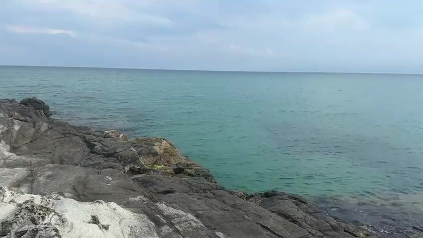 The video shows a rocky coastline with clear turquoise water. The sky is partly cloudy with some sunlight filtering through. The camera pans from left to right, revealing a wider view of the coastline, including a sandy beach in the distance and rolling hills covered in vegetation. There are no people or objects visible in the video. The waves are gentle, and the overall atmosphere is serene and natural.