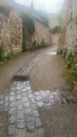 The video presents a walk through a narrow, old-world street in Saint-Leu-d'Esserent, France. The scene is dominated by tall, ancient stone walls on both sides, some covered with patches of greenery and ivy. The ground is a mix of asphalt and historic cobblestones, with a prominent stone-lined drainage channel running down the center of the path, leading to a metal grate in the foreground. The street appears damp, suggesting recent rain or morning dew.

The camera begins by looking down the street, then pans upwards along a weathered stone wall. A traditional, dark-colored street lamp with a square housing is mounted on this wall. A warm, yellowish-orange light is reflected on the top edge of the wall, possibly from an unseen light source or a fleeting moment of sunlight breaking through the overcast sky, which appears bright but diffused. The sky is largely grey and cloudy.

The pan then returns to street level, showing the continued path of cobblestones and the winding nature of the alley. Distant buildings and trees are visible beyond the stone structures, contributing to the tranquil and historic atmosphere. There are no people visible, and the overall impression is one of quiet solitude in a charming, ancient setting.
