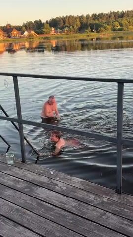 Two men are in the water near a wooden dock with metal railings and a ladder. One man is submerged up to his chest and appears to be treading water. The other man is swimming towards the ladder. A glass bottle is on the dock. In the background, there is a lake, trees, and buildings. The sky is clear and the sun is low in the sky, suggesting it is either sunrise or sunset. The scene appears to be a recreational area.