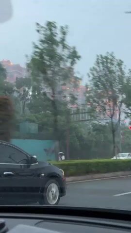 From inside a car on a rainy day, a black sedan is observed on a road. The windshield wipers are in motion. In the background, there's a large building with a distinctive, multi-tiered, ornate design illuminated with orange lights. Trees and greenery surround the building. Other cars are also visible on the road. The license plate on the sedan is partially visible, showing "JC531."