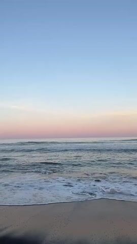 A drone's eye view captures a tranquil beach scene at sunrise or sunset. The camera slowly pans left, revealing a vast expanse of the ocean meeting a dark, sandy shore. Gentle waves crash onto the beach, leaving foamy trails. The sky is a gradient of soft blues and pinks, with a few wispy clouds. In the distance, a faint outline of mountains can be seen on the horizon. The overall atmosphere is peaceful and serene, with no people or significant objects present to distract from the natural beauty of the environment.