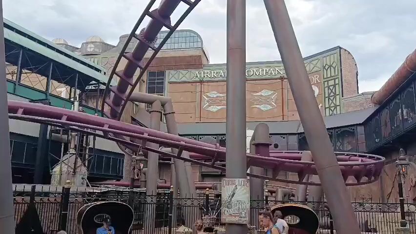 A roller coaster train, filled with riders, is captured mid-ride on a section of track that appears to be going downwards. The roller coaster is predominantly purple with dark accents, and the train is a dark color. The setting is an amusement park, with a building in the background featuring large lettering that reads "AIRSHIP COMPANY". The building's architecture suggests a steampunk or industrial theme. Several people are visible in the foreground, some appearing to be waiting in line for the ride, while others are observing. The sky is overcast, indicating a cloudy day. The entire scene is shot from a static position, offering a clear view of the roller coaster in motion.