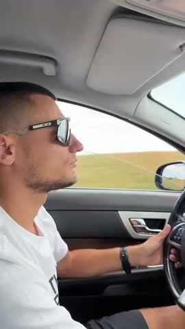 The video begins with a close-up shot of a man wearing sunglasses and a white t-shirt, driving a car. He is looking out the window to his left. The camera then pans to the interior of the car, showing the steering wheel, dashboard, and rearview mirror. The view through the windshield reveals a long, empty road stretching into the distance, bordered by dry, grassy fields. In the far distance, mountains can be seen under a partly cloudy sky. The camera continues to move, showing the passenger side of the car and the passing landscape. The driver's hand rests on the steering wheel. The video ends with a shot of the driver and the exterior scenery through the passenger window. The overall impression is a scenic drive through a rural, possibly mountainous area.