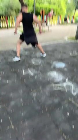 A man in athletic wear is playing hopscotch in a park. He is in the process of hopping between numbered squares drawn on the ground with chalk. In the background, there are benches, a colorful fence, trees, and other people. The man appears to be enjoying himself as he plays the game.