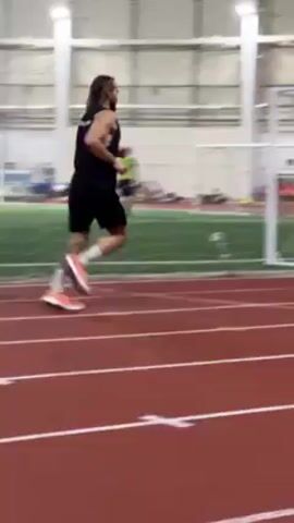 A man with long hair, wearing a black tank top, black shorts, and orange and white running shoes, runs along a red track indoors. The track has white lane markings and is next to a green field. There are white soccer goals on the field. Spectators are visible in the background. The indoor facility has a high ceiling with exposed white support beams and overhead lighting. A second man is seen walking ahead of the runner on the track.