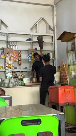 Two young men in black shirts are in a shop. The man on the left is holding a long, dark fabric bag, possibly a tea strainer, high in the air with his right arm. He is standing behind a counter with various items on it, including cups, glasses, and what appear to be packaged goods on shelves behind him. The man on the right is standing to the left of the first man and appears to be holding a cup. They are both facing towards the right side of the frame. The environment looks like a small shop or food stall with shelves stocked with items. There is a red cooler box to the right of the counter. The video is a short clip showing this brief interaction.