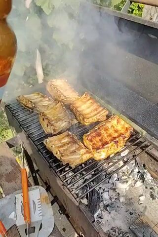 The video shows a close-up of several racks of ribs grilling over hot coals. Smoke billows around the ribs, and the grill is in the center of the frame. The background is blurred and appears to be an outdoor setting with greenery. The bottom of the frame shows a hand holding a bottle of what looks like barbecue sauce. The ribs appear to be seasoned and are starting to char. The overall scene depicts a casual outdoor cooking event.