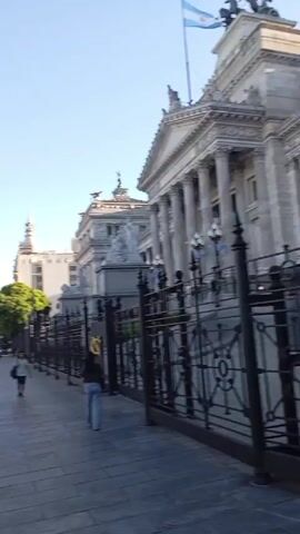 The video captures a cityscape on a sunny day. Cars are lined up in traffic on the left side of the street, while pedestrians walk along the paved sidewalk to the right. A large, ornate building with decorative statues is visible to the right, partially obscured by a metal fence. There is a tall tower in the distance. People are walking across a crosswalk, while cars are stopped at a red light. The scene is typical of a busy urban environment.