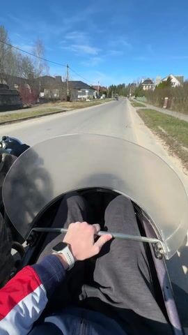 A first-person perspective video shows a person riding in the sidecar of a motorcycle on a sunny day. The passenger holds onto a metal bar within the sidecar, wearing black pants, a red and blue jacket, and a white smartwatch. The driver, wearing a camouflage jacket and helmet, steers the motorcycle down a residential street. Houses are visible on both sides of the road, and a car can be seen in the distance. The sky is blue with scattered clouds.