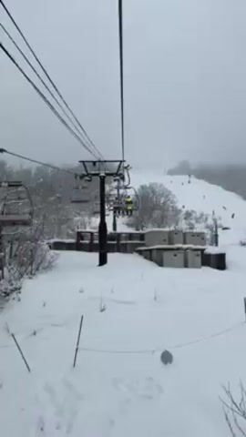 A ski lift carries passengers in a snowy, overcast environment. The lift's black support poles anchor the overhead cables. Chairs, some red and others carrying skiers, move along the cables. The skiers are wearing various attire, including a bright yellow jacket. The ground is covered in deep snow, with scattered vegetation poking through. A low wall runs along the bottom of the frame, interspersed with concrete blocks. Skiers are visible on the snowy hill in the distance. It is snowing lightly.