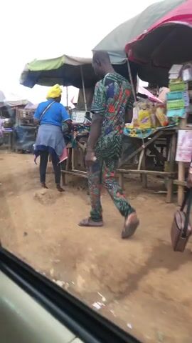 A blurry video shows a busy outdoor market scene. A man in a colorful patterned outfit walks towards the camera. In the background, a woman in a blue shirt and yellow headscarf walks away. Further in the background, a person in a striped shirt carries a large black bag over their head, walking away from the camera. Stalls with awnings are set up along a dirt path. Several motorcycles are parked in a row. The sky is overcast.