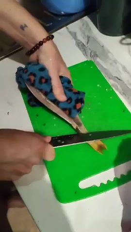 A pair of hands prepares a fish on a green cutting board. One hand, adorned with a beaded bracelet, holds a blue and orange leopard-print cloth over the fish, while the other hand grips a knife. The fish lies on its side, its body elongated with scales visible. The cutting board is placed on a white countertop, with a stainless steel sink and a blue bowl in the background. The scene appears to be indoors, likely a kitchen, given the sink and countertop. The lighting suggests it could be daytime, but no definitive clues about the time of day or weather are present. There is no visible text in the media. The activity is the preparation of a fish, possibly for cooking, as indicated by the knife and cutting board. The focus is on the methodical process of handling the fish, suggesting a practical, domestic setting.