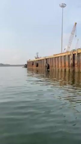 The video shows a waterfront scene with a rusty, metal pier. The camera moves steadily through the water, alongside the pier. The pier has several black, round fenders along its side. On the pier, there are cranes and stacks of shipping containers. A tall light pole is also visible. The water is greenish-gray and reflects the pier. The sky is bright with a few clouds.