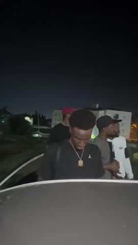 The video, shot at night, appears to be taken from the roof of a moving vehicle. Four men are seen in the foreground. The man closest to the camera wears a black shirt and a gold chain.  Behind him, another man wears a baseball cap and a gray shirt. To his right, a man in a white and black hoodie is visible. The fourth man, in a red cap, is mostly out of focus. In the background are buildings and streetlights, blurred by the motion of the vehicle. The camera shakes due to the movement, and light streaks indicate the vehicle's speed.