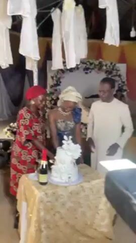 Here's a description of the video:

A wedding scene shows a bride in a blue and gold dress and headwrap, standing at a table with a tiered white cake adorned with white flowers.  On her left is a woman in a red patterned dress and headwrap. To the bride's right is a man in a long-sleeved beige shirt. They appear to be preparing to cut the cake, with a bottle of champagne visible on the table. The table is covered with a gold sequined tablecloth.

Behind them is a floral arch decorated with white and purple flowers, set against a backdrop of draped fabric. The ceiling is decorated with white cloth hangings. A camera on a tripod is visible in the foreground.  The lighting appears to be from overhead sources.