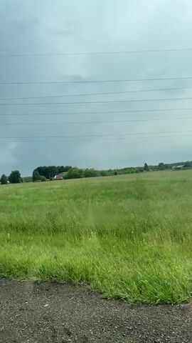 A video taken from a moving vehicle shows a rural landscape. A power pole and lines are visible on the left side of the frame. A wide, green grassy field stretches out before the viewer, dotted with trees in the distance. Several buildings, possibly houses or farm structures, are scattered across the field. The sky is overcast and appears a muted gray. A blue sign with white icons indicating a bed (for lodging) and cutlery with a fork and knife (for dining), along with the number "300m," is briefly visible on a pole in the field. The ground in the foreground appears to be a dirt or gravel road.
