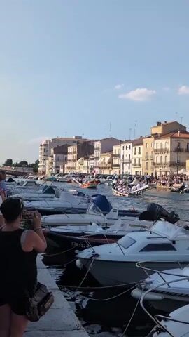 The video captures a waterside scene with numerous small boats docked along a quay. A woman in the foreground, with her back to the camera, is holding a phone up, seemingly taking a picture of the activity in the distance. Beyond the docked boats, several larger vessels are present. One boat, with a crew dressed in white, is actively engaged in rowing, moving through the water. Along the shore in the background, a line of multi-story buildings with light-colored facades and balconies overlooks the water. A crowd of spectators is gathered along the waterfront, watching the rowing event. Cars are parked along the street behind the buildings. The sky is clear and blue with a few scattered clouds, indicating fair weather. The lighting suggests it is daytime, likely afternoon.