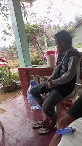 The video shows a man sitting on a white plastic chair on a covered porch. He is wearing a black and gray long-sleeved shirt, blue jeans, and black flip-flops. He is holding a black smartphone in his left hand and appears to be smoking. To his left is a large bottle of water and a red and white cooler. In the background, there is a building with pink flowers growing on it, a wooden fence, and trees. Another person's legs and feet, wearing flip-flops, are visible in the foreground on the right. Another man wearing a red t-shirt and shorts, and a baseball cap is sitting on a similar chair to the left of the frame, smoking. He is holding a book or notebook in his lap. A motorcycle is parked in the background to his left. The ground is tiled on the porch and dirt outside. The sky is overcast.