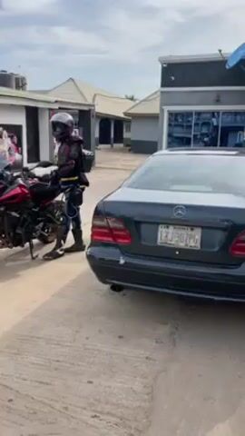 The video shows two motorcycles parked outside a building. One is red and black, and a person in riding gear stands beside it. The other motorcycle is blue, white, and silver. The surrounding area seems to be a parking lot in front of some buildings. There are advertisements or posters displayed on one of the buildings. A black car is parked near the first motorcycle. A person in a light-colored riding jacket walks toward the second motorcycle.