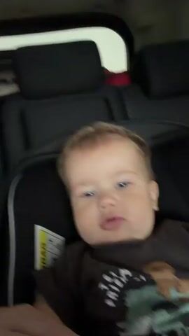 A baby is sitting in a car seat, looking at the camera and making a face. The baby is wearing a dark t-shirt with a graphic design. The background shows the interior of a car with black headrests. The video is shaky and the focus shifts.