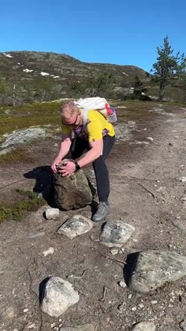 A man wearing a yellow shirt, dark pants, gray hiking shoes, and a backpack, is bent over, inspecting a large gray rock. He is on a dirt and rocky path in a mountainous area with sparse vegetation, under a bright blue sky. The rock he is examining is surrounded by smaller rocks. In the background, there are hills with patches of snow, scattered trees, and low-lying scrub. The man's focus is on the rock, and he appears to be closely examining its surface.